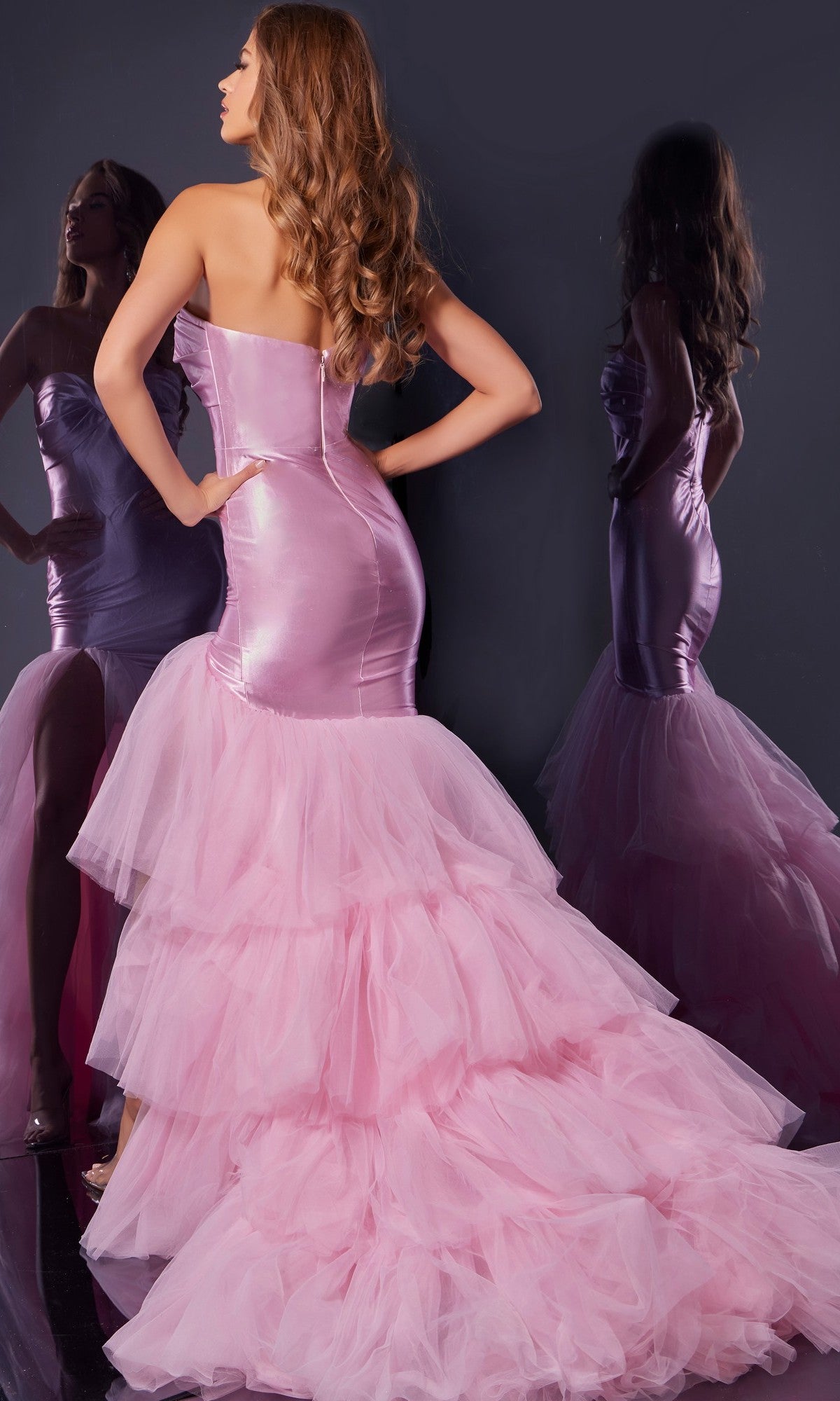 A woman stands before a mirror in the stunning Jovani D5315, a strapless ruffled mermaid prom gown with a dramatic tulle train, showcasing the elegant pink dress from behind.